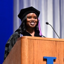 woman wearing graduation regalia, speaking into a podium. 