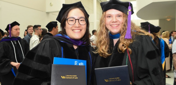 Two recent graduates in commencement regalia holding up their degrees, smiling. 