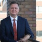 Man wearing blue suit, red tie, smiling, standing outside next to a blue column. 