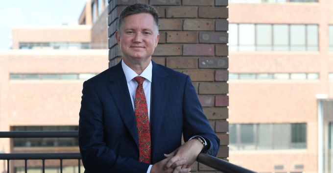 Man wearing blue suit, red tie, smiling, standing outside next to a blue column. 