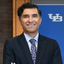 man wearing business suit, smiling, standing in front of a UB sign. 