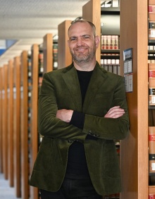 Man standing, leaning against a library bookshelf, arms folded, smiling. 