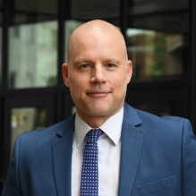 Man wearing blue suit, standing outside a building, smiling.