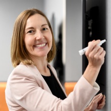 Woman smiling, writing on a white board in a classroom.