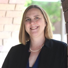 Woman wearing black jacket, standing outside, next to a brick wall.