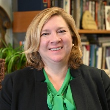 Woman wearing black sweater over green shirt, smiling, standing in her office. 