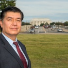 man wearing grety business suit standingn outside on a grassy lawn in Washington, D.C., Lincoln memorial building in the distance. 