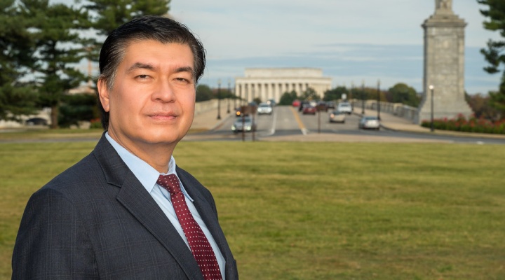 man wearing grety business suit standingn outside on a grassy lawn in Washington, D.C., Lincoln memorial building in the distance. 