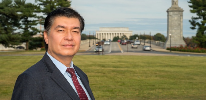 man wearing grety business suit standingn outside on a grassy lawn in Washington, D.C., Lincoln memorial building in the distance. 