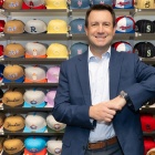 Man in a suit standing casually, smiling, with several sales racks of colorful baseball caps behind him. 