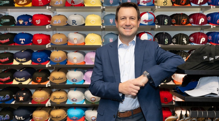 Man in a suit standing casually, smiling, with several sales racks of colorful baseball caps behind him. 