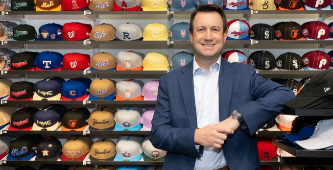 Man in a suit standing casually, smiling, with several sales racks of colorful baseball caps behind him. 