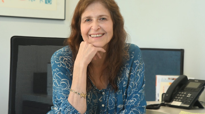 Woman wearing blue dress, sitting at her desk, smiling. 