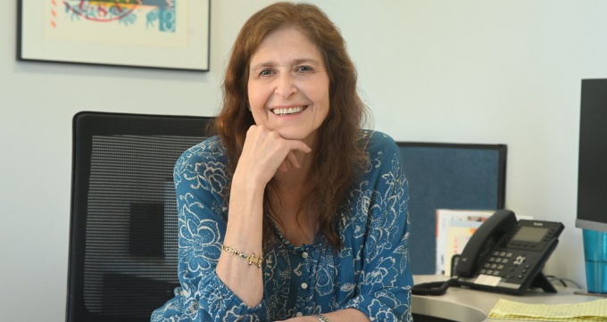 Woman wearing blue dress, sitting at her desk, smiling. 