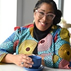 Woman wearing glasses, colorful jacket, sitting in a cafe with a coffee cup, smiling. 