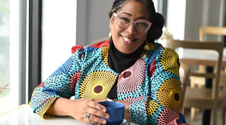 Woman wearing glasses, colorful jacket, sitting in a cafe with a coffee cup, smiling. 