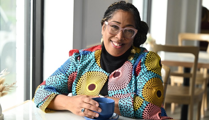 Woman wearing glasses, colorful jacket, sitting in a cafe with a coffee cup, smiling. 