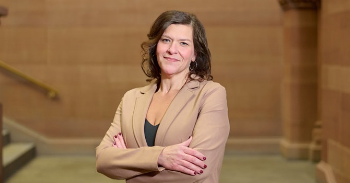 Woman wearing beige suit, arms crossed, smiling, standing in a large stairwell. 