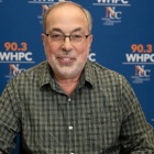 Man sitting at a table, smiling, blue wall behind him says 90.3 WHPC. 