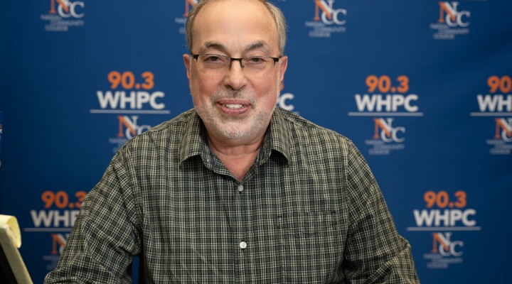 Man sitting at a table, smiling, blue wall behind him says 90.3 WHPC. 