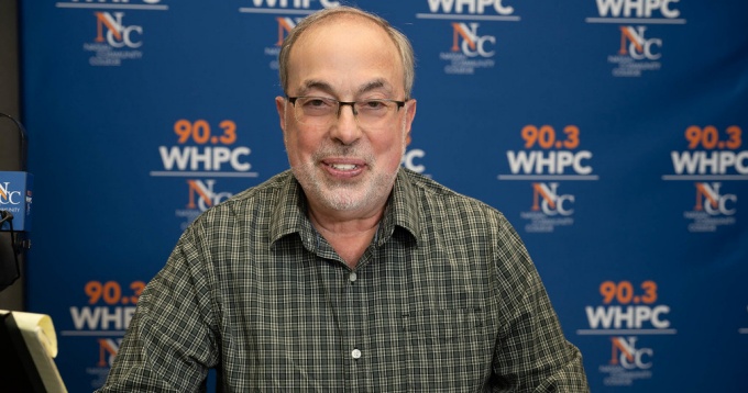 Man sitting at a table, smiling, blue wall behind him says 90.3 WHPC. 