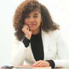 Woman wearing white jacket, black top, sitting at a table with notepads, in a white room. 