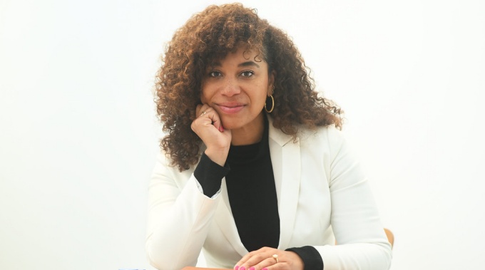 Woman wearing white jacket, black top, sitting at a table with notepads, in a white room. 