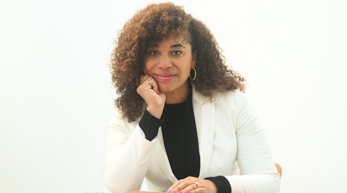 Woman wearing white jacket, black top, sitting at a table with notepads, in a white room.
