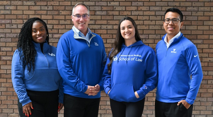 Four individuals standing in front of a brick wall, all wearing University at Buffalo School of Law apparel in shades of blue. Two are in quarter-zip pullovers with UB branding, one in a hoodie with “University at Buffalo School of Law” printed on the front, and one in a crewneck sweatshirt with the UB logo. They are posed side by side, facing forward.