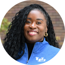 Close-up of a person wearing a royal blue University at Buffalo zip-up jacket with the white “UB” logo on the left side. The jacket is partially zipped, revealing a black shirt underneath. The person has long, curly black hair and is wearing gold hoop earrings. The background is softly blurred with earthy tones.