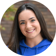 Close-up of a person wearing a bright blue hoodie with the words “University at Buffalo School of Law” printed in white on the front. The hoodie has drawstrings visible near the neckline. The background shows a brick wall with some greenery and autumn-colored leaves.