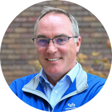 Close-up of a person wearing a blue University at Buffalo jacket with a light gray collar and a white button-down shirt underneath. The jacket features the white “UB” logo and the words “University at Buffalo” on the left side. The background shows a brick wall with some greenery and autumn-colored leaves.