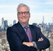 Person wearing a dark navy suit with a light blue dress shirt and a patterned purple tie, standing with arms crossed on a rooftop or balcony overlooking a city skyline. The background features numerous high-rise buildings under a partly cloudy sky.