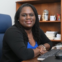 Person seated at a wooden desk in an office setting, wearing a blue top with a black cardigan. The desk has a computer keyboard, phone, and office supplies. Behind the person is a wooden bookshelf with books, framed photos, and decorative items.