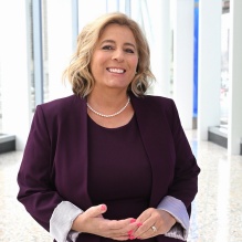 Person wearing a deep purple dress and matching blazer with light-colored cuffs, accessorized with a pearl necklace and pink nail polish. The individual is standing in a bright, modern interior space with large floor-to-ceiling windows and a terrazzo floor.
