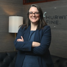 Person standing with arms crossed in a modern office setting. The individual is wearing a navy blazer over a patterned blue top. Behind them is a wall with a mounted sign that reads “Hurwitz Fine P.C.” and a floor lamp next to a black tufted leather sofa.