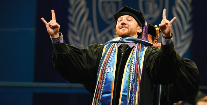 Graduate wearing a black cap and gown adorned with multiple colorful honor cords and stoles raises both hands in a celebratory gesture during a commencement ceremony. A large blue backdrop behind shows the University at Buffalo School of Law emblem featuring a buffalo and shield design.