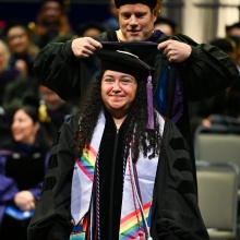 Graduate in black academic regalia with a purple tassel and a colorful stole featuring rainbow stripes, standing while another person places a hood over their shoulders. Several other individuals in academic attire are seated in the background.