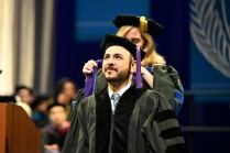 A person in academic regalia is being hooded during a graduation ceremony. Another individual, also in academic regalia, places a purple graduation hood over the person’s shoulders. The setting includes a stage with a podium on the left, several people seated in the background, and a large blue banner featuring a laurel design.