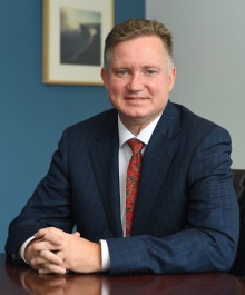 Person seated at a wooden table wearing a dark blue checkered suit jacket, white dress shirt, and a red patterned tie. Hands are clasped on the table. Behind is a blue wall with a framed picture hanging on it.