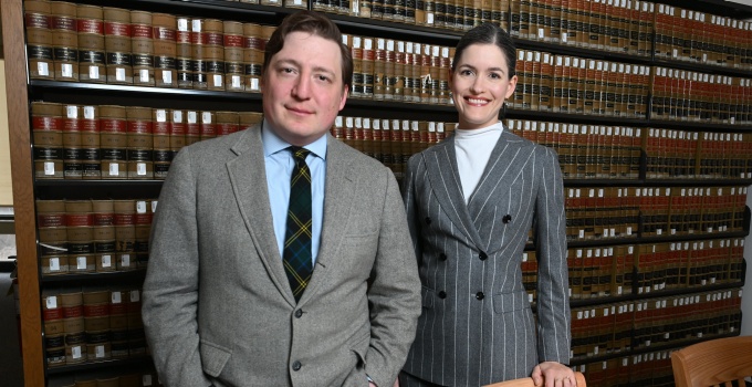 Two people wearing business attire stand in front of shelves filled with rows of legal books in a law library. A wooden table with chairs is visible in the foreground.