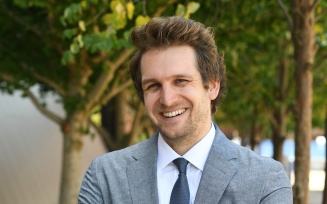 Person wearing a light gray suit with a white dress shirt and dark tie stands outdoors with arms crossed. The background shows green trees, shrubs, and part of a building wall.