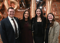Four people standing together in an indoor event space with exposed brick walls and large metallic balloon letters in the background. They are dressed in formal attire: one in a dark suit with a tie, two in black outfits, and one in a light plaid blazer.