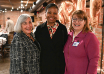 Three people standing close together in a warmly lit indoor space with exposed brick walls and metallic balloon decorations in the background. One person is wearing a patterned jacket, another is in a black outfit with a floral top underneath, and the third is in a bright pink zip-up jacket with a visible name tag.
