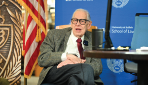Person seated indoors wearing a gray tweed blazer over a white shirt with a red tie. An American flag is visible on the left, and a blue backdrop with the University at Buffalo School of Law logo is in the background. A microphone and part of a table appear in the foreground.