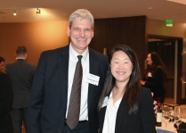 Two people standing together at an indoor event. One person is wearing a dark suit with a white dress shirt and patterned tie, and the other is wearing a black top with a white blazer. Both have visible name tags, and a table with wine bottles is in the background.
