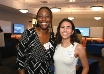 Two people standing together at an indoor event space with round ceiling lights and presentation screens in the background. One person is wearing a black-and-white patterned dress with a name tag, and the other is wearing a sleeveless white top with a name tag.