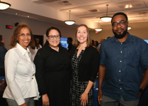 Four people standing together at an indoor event space with round ceiling lights and a crowd in the background. One person is wearing a white wrap-style blouse, another is in a black dress, the third is in a black-and-white patterned dress with a black cardigan, and the fourth is in a dark blue button-up shirt.
