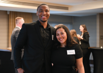 Two people standing together at an indoor event. Both are wearing black outfits; one is in a black suit with a collared shirt, and the other is in a short-sleeved black dress with a visible name tag. Other attendees and cocktail tables are visible in the background.