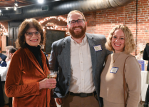 Three people standing together at an indoor event with exposed brick walls and string lights. One person is wearing a rust-colored velvet jacket and holding a drink, another is in a gray blazer over a white shirt, and the third is in a beige top with a necklace. Two visible name tags are worn on the gray blazer and beige top.